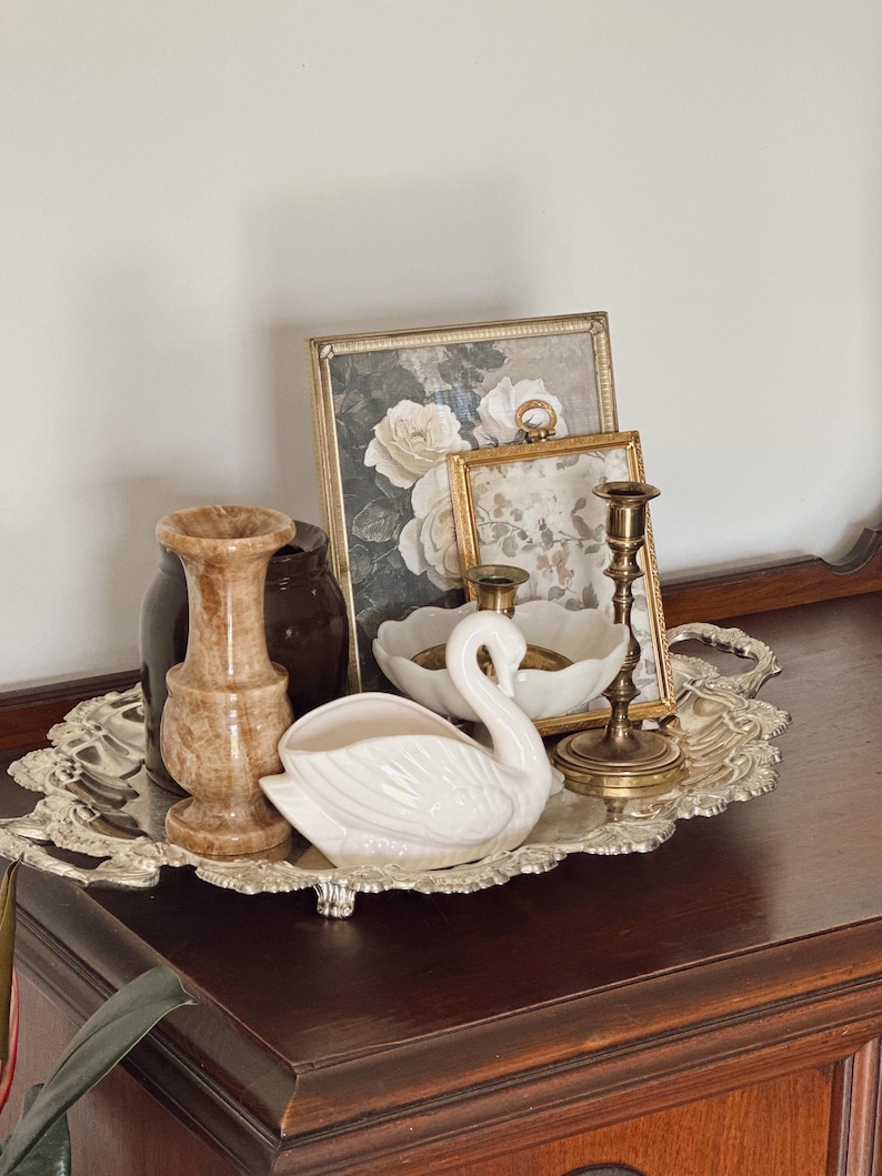May include: A decorative silver tray holds a collection of vintage items. A white ceramic swan, two framed floral prints, a marble-like vase, a dark brown jar, a white bowl, and two brass candlesticks are arranged on the tray.