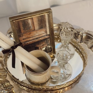 May include: A decorative silver tray with a mirrored surface holds a framed picture of a piano, a mug with two white candles, and two clear glass candle holders. A dark brown ribbon ties the candles together.