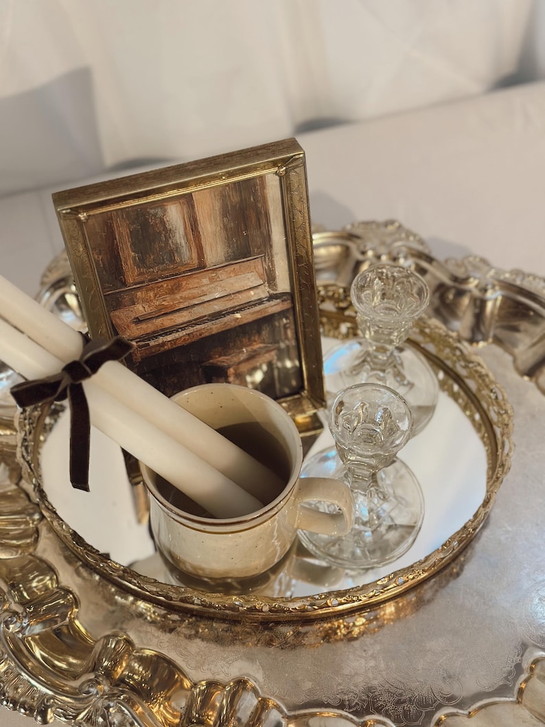 May include: A decorative silver tray with a mirrored surface holds a framed picture of a piano, a mug with two white candles, and two clear glass candle holders. A dark brown ribbon ties the candles together.