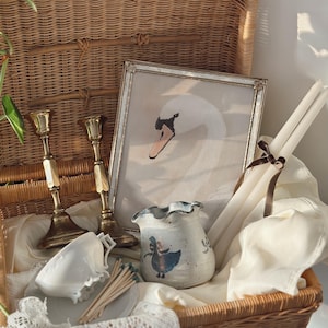 May include: A wicker basket filled with vintage items. Includes a framed swan painting, brass candlesticks, a ceramic vase with a blue illustration, white candles, a teacup, and matches. The basket is lined with white fabric and lace.