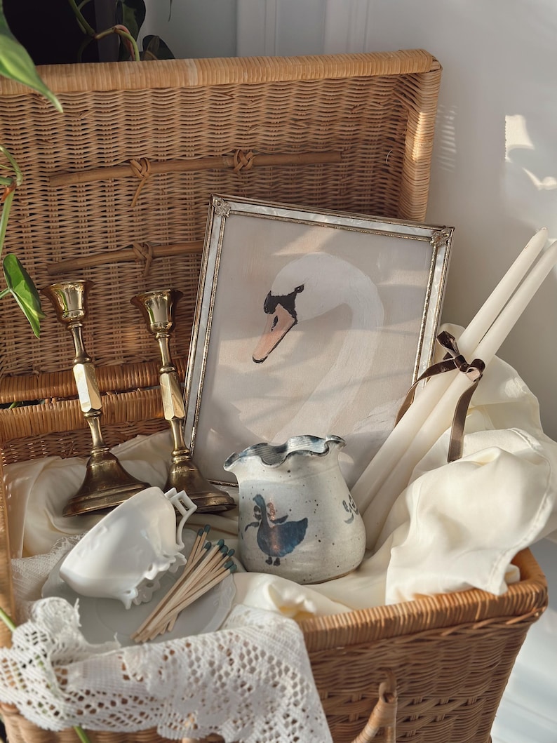 May include: A wicker basket filled with vintage items. Includes a framed swan painting, brass candlesticks, a ceramic vase with a blue illustration, white candles, a teacup, and matches. The basket is lined with white fabric and lace.
