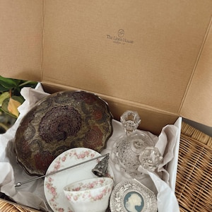 May include: An open cardboard box filled with vintage items. Includes a decorative metal bowl, a floral teacup and saucer, a crystal perfume bottle, a silver compact, and a decorative bell. The box is lined with white tissue paper.