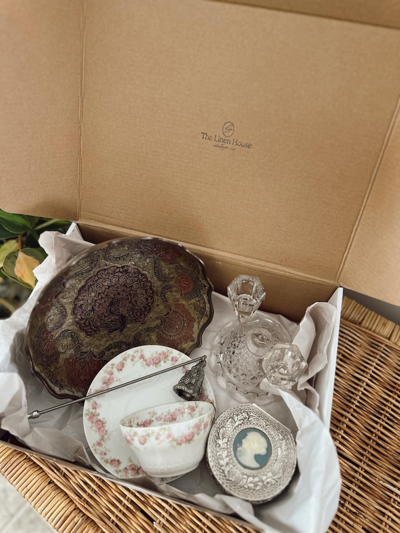 May include: An open cardboard box filled with vintage items. Includes a decorative metal bowl, a floral teacup and saucer, a crystal perfume bottle, a silver compact, and a decorative bell. The box is lined with white tissue paper.
