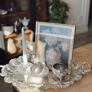 May include: A silver-plated serving tray with ornate edges holds a framed picture, a candle, a small dish with a lid, and a creamer. The picture features a floral still life. The tray sits on a wooden table.