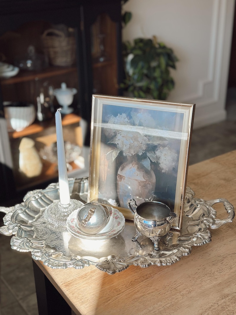 May include: A silver-plated serving tray with ornate edges holds a framed picture, a candle, a small dish with a lid, and a creamer. The picture features a floral still life. The tray sits on a wooden table.