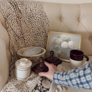 May include: A still life arrangement featuring a framed floral print, a brown and white mug, a small ceramic jar with blue stripes, and two wooden candle holders. A patterned throw and a decorative bowl complete the scene.