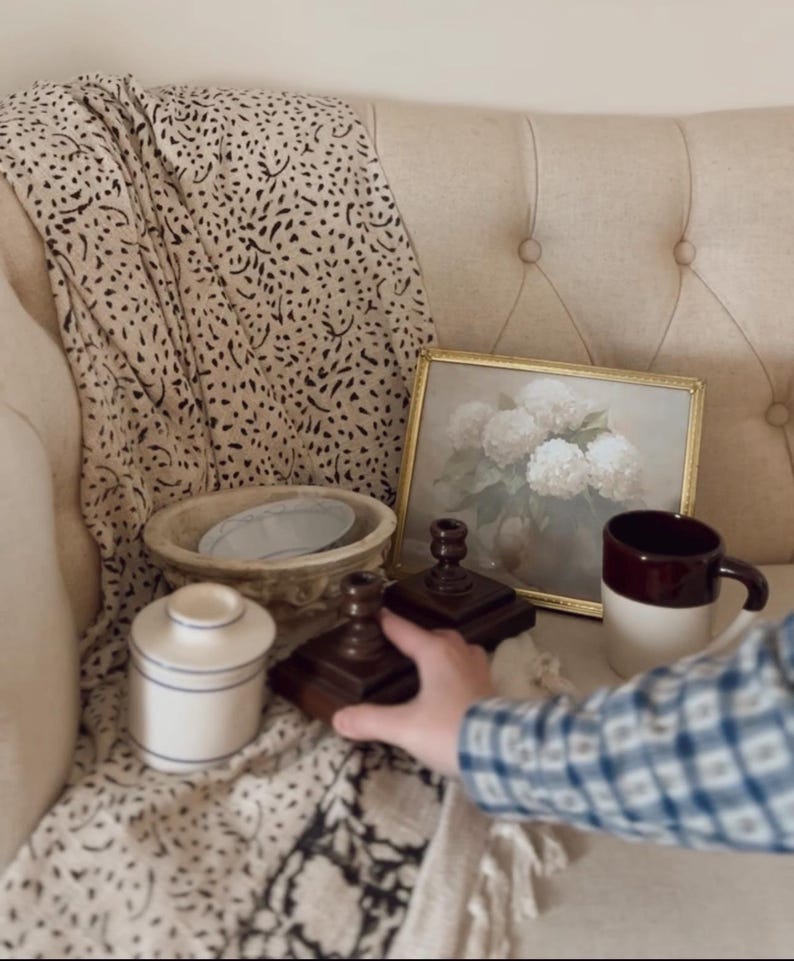 May include: A still life arrangement featuring a framed floral print, a brown and white mug, a small ceramic jar with blue stripes, and two wooden candle holders. A patterned throw and a decorative bowl complete the scene.