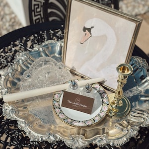 May include: A silver serving tray holds a framed swan painting, a candle, a floral plate with salt and pepper shakers, and a gold candlestick. A business card for "The Linen House" rests on the plate. The tray sits on a black, ornate table.