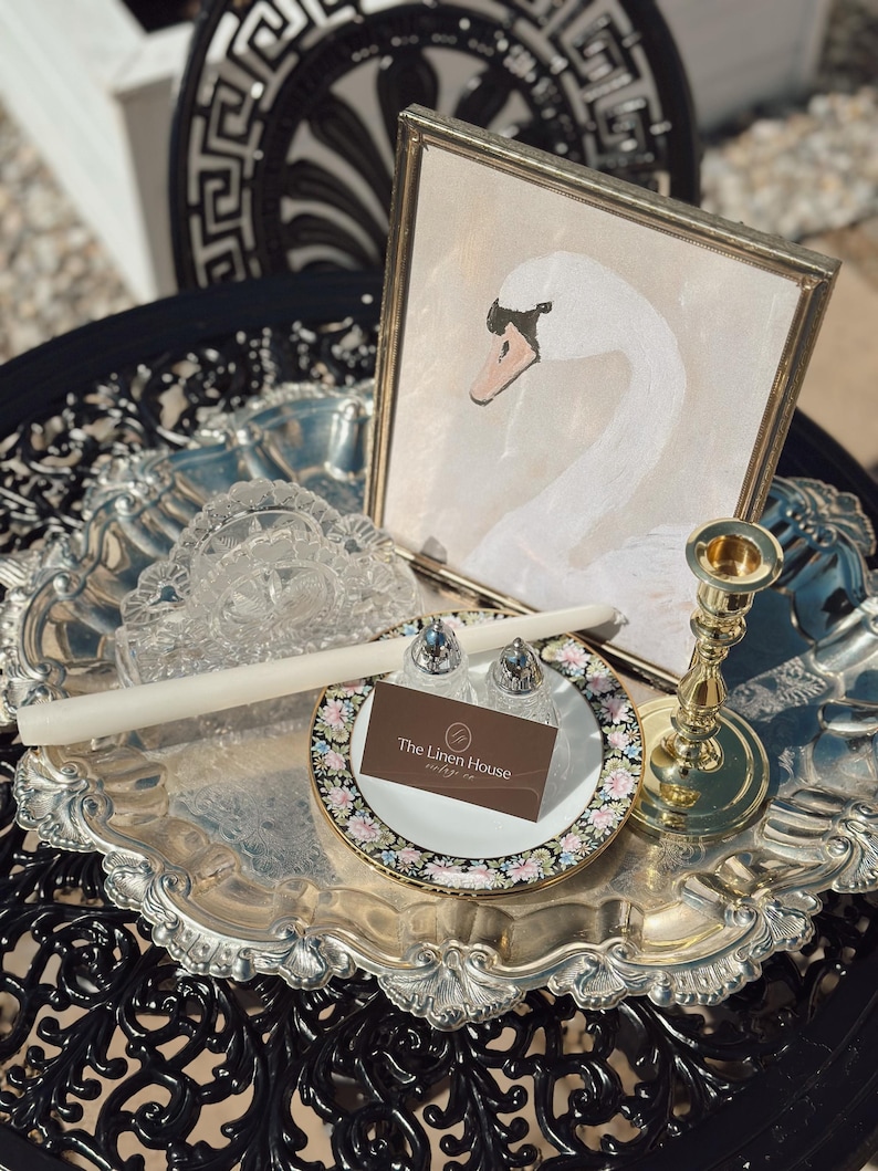 May include: A silver serving tray holds a framed swan painting, a candle, a floral plate with salt and pepper shakers, and a gold candlestick. A business card for "The Linen House" rests on the plate. The tray sits on a black, ornate table.