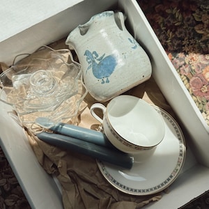 May include: A collection of vintage items in a cardboard box. Includes a ceramic pitcher with a blue bird design, a glass candle holder, two blue candles, a teacup, and a saucer with a blue floral border. The items are nestled in brown packing paper.