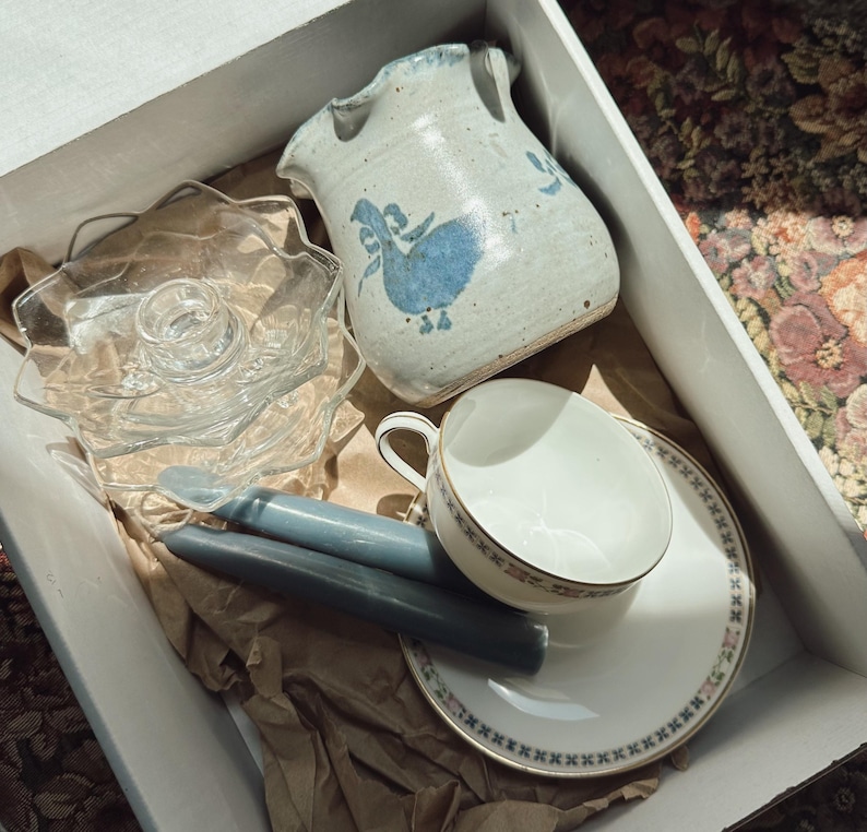 May include: A collection of vintage items in a cardboard box. Includes a ceramic pitcher with a blue bird design, a glass candle holder, two blue candles, a teacup, and a saucer with a blue floral border. The items are nestled in brown packing paper.