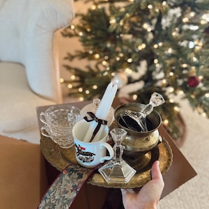 May include: A collection of vintage items, including a brass tray, teacups, a mug with a horse illustration, candles, and a decorative ribbon. The items are arranged in a cardboard box, with a Christmas tree in the background.