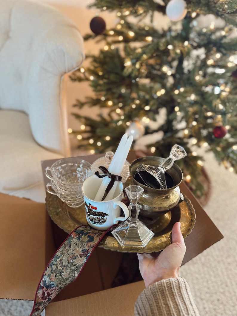 May include: A collection of vintage items, including a brass tray, teacups, a mug with a horse illustration, candles, and a decorative ribbon. The items are arranged in a cardboard box, with a Christmas tree in the background.