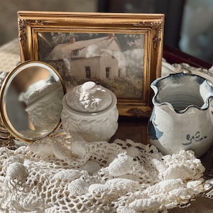 May include: An antique still life arrangement featuring a gold-framed picture of a house, a small white lidded jar, a round mirror, and a blue and white ceramic vase. A white lace doily is draped across the objects.