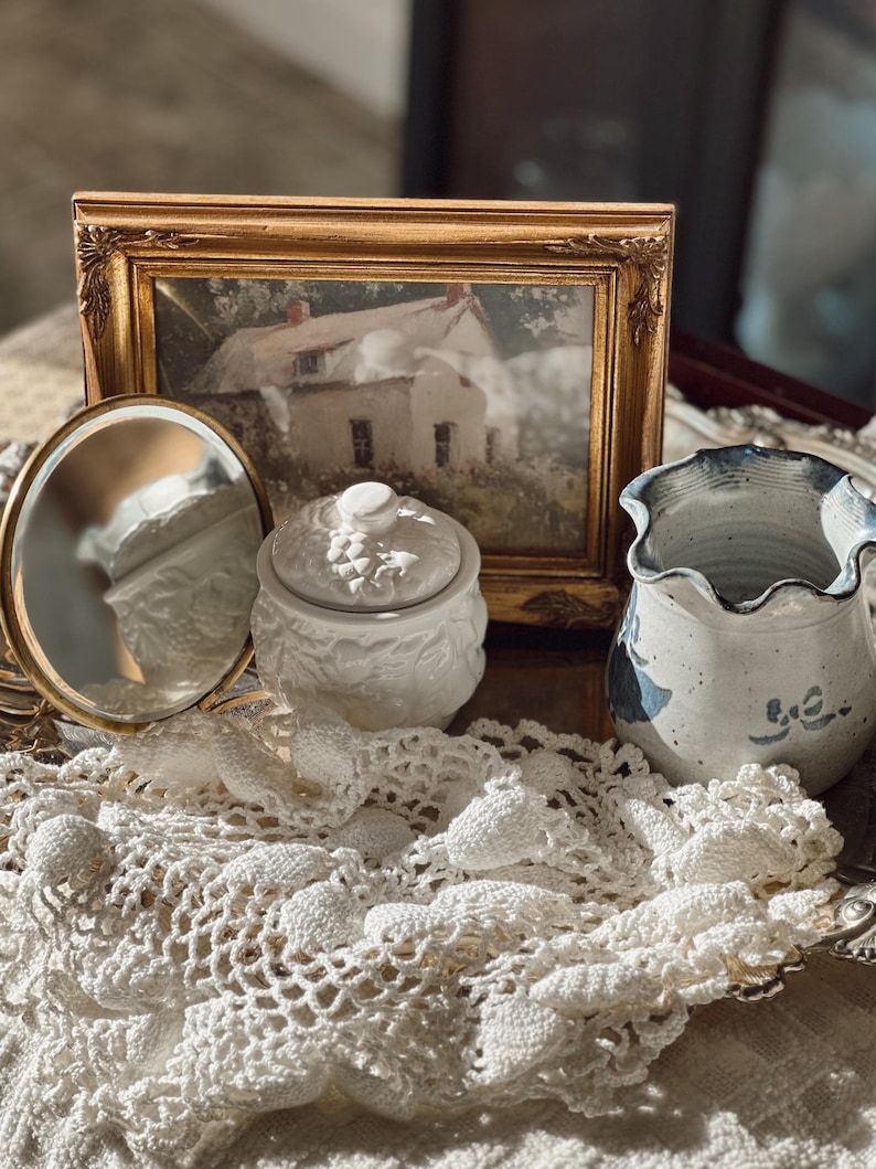 May include: An antique still life arrangement featuring a gold-framed picture of a house, a small white lidded jar, a round mirror, and a blue and white ceramic vase. A white lace doily is draped across the objects.