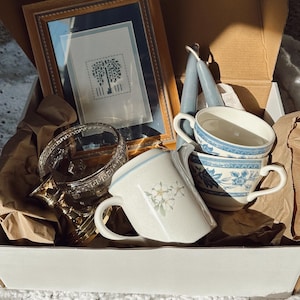 May include: A box filled with vintage items. Includes a framed cross-stitch picture, two blue taper candles, a glass and gold-toned decorative piece, and several teacups with floral and blue patterns. The box is lined with brown paper.
