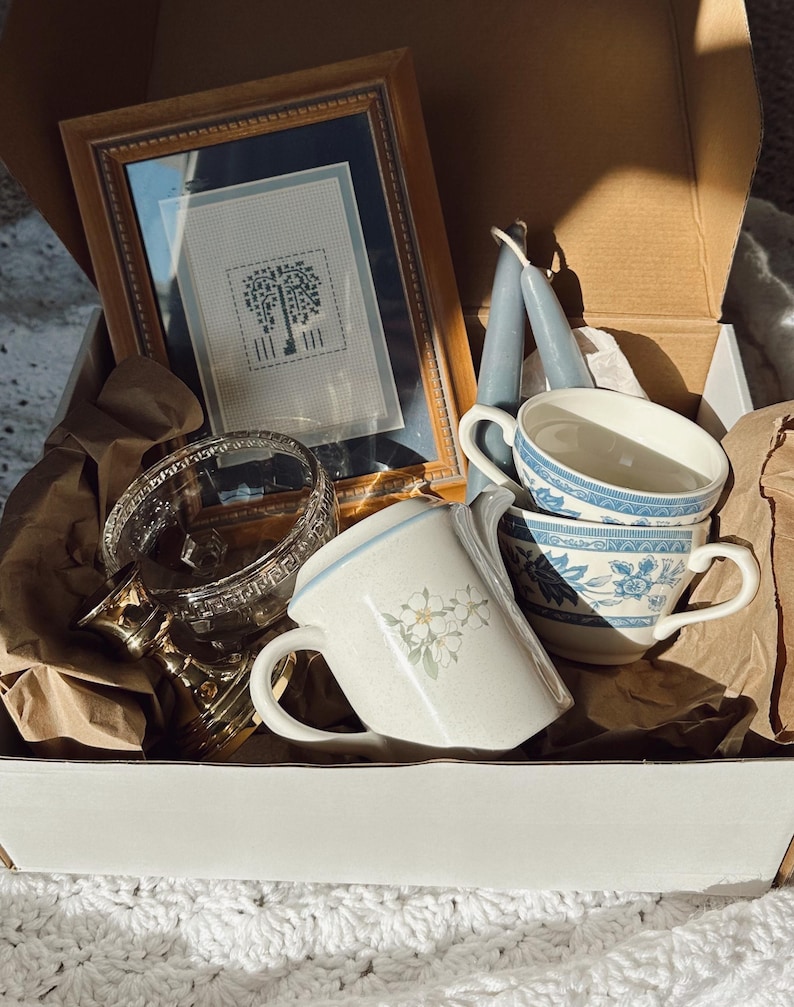 May include: A box filled with vintage items. Includes a framed cross-stitch picture, two blue taper candles, a glass and gold-toned decorative piece, and several teacups with floral and blue patterns. The box is lined with brown paper.
