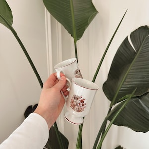 May include: Two white ceramic mugs with brown floral designs and gold trim. The mugs have a footed base and are held in a hand.