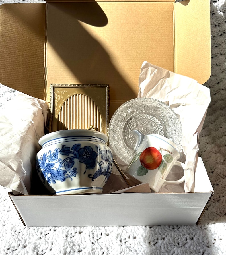 May include: A gift box containing a blue and white floral ceramic pot, a small gold picture frame, a clear glass plate, and a white teacup with a red apple design. The items are nestled in white packing paper.