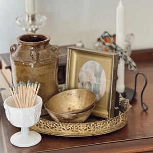 May include: A still life arrangement featuring a brown ceramic jar, a white and gold framed picture, two small brass bowls, and a white decorative match holder filled with matches. A decorative gold tray holds the bowls and picture.