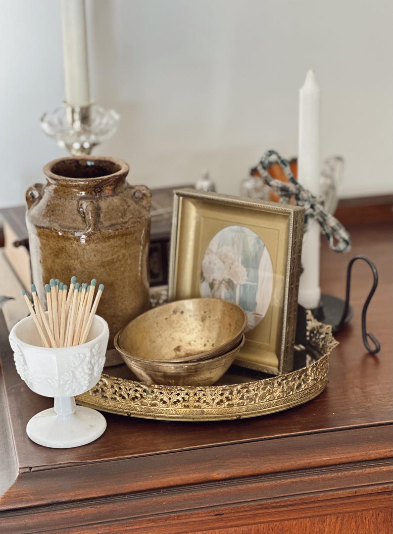May include: A still life arrangement featuring a brown ceramic jar, a white and gold framed picture, two small brass bowls, and a white decorative match holder filled with matches. A decorative gold tray holds the bowls and picture.