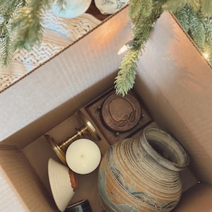 May include: A cardboard box filled with various decorative items. The box contains a ceramic vase with blue and brown patterns, a wooden coaster, a brass candle holder, a white candle, and other small objects. The items are arranged inside the box.