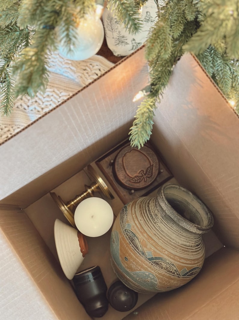 May include: A cardboard box filled with various decorative items. The box contains a ceramic vase with blue and brown patterns, a wooden coaster, a brass candle holder, a white candle, and other small objects. The items are arranged inside the box.