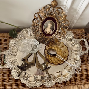 May include: A silver-colored, ornate tray holds a collection of vintage items. These include a gold-framed portrait, amber-colored glass bowl, silver serving tongs, two brass candle holders, and a clear glass dish. The tray sits on a woven wicker surface.