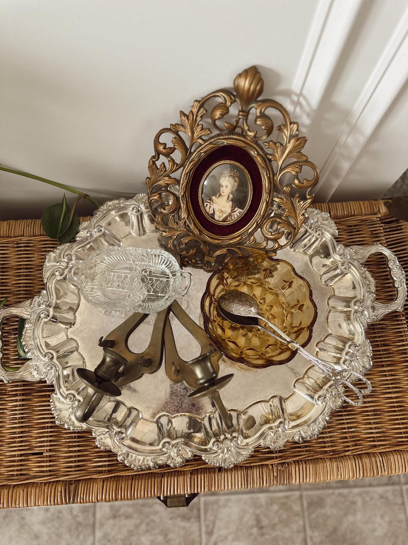 May include: A silver-colored, ornate tray holds a collection of vintage items. These include a gold-framed portrait, amber-colored glass bowl, silver serving tongs, two brass candle holders, and a clear glass dish. The tray sits on a woven wicker surface.