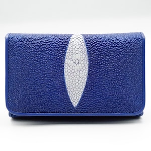 Puede incluir: Una cartera rectangular con un exterior azul texturizado y una incrustación ovalada blanca alargada en el centro. La superficie tiene una apariencia granulada y rugosa característica de la piel de raya, con bordes definidos.