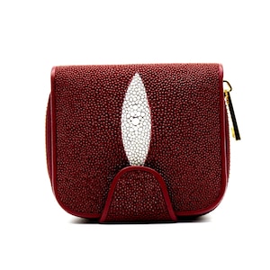 May include: A burgundy leather wallet with a white stingray design. The wallet has a textured surface and a gold zip. The design features a central white oval shape with a dotted pattern, adding a unique touch to the accessory.