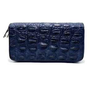 Puede incluir: Una cartera rectangular de color azul marino con un patrón texturizado en relieve de piel de cocodrilo en toda la superficie exterior. La cartera está asegurada con un tirador de cremallera de metal plateado visible en el lado izquierdo.