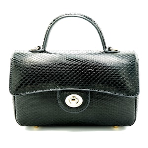 Könnte beinhalten: Eine schwarze Handtasche mit einem strukturierten, schlangenartigen Muster. Die Tasche hat einen oberen Griff und einen Schnallenverschluss. Die Hardware ist goldfarben. Die Tasche ist rechteckig mit abgerundeten Ecken und kleinen goldenen Füßen.