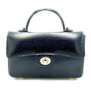 Könnte beinhalten: Eine marineblaue Handtasche mit einem strukturierten Wabenmuster. Die Tasche hat einen oberen Griff, einen goldfarbenen Verschluss und kleine goldene Füße. Das Design ist strukturiert und elegant, passend für verschiedene Anlässe.