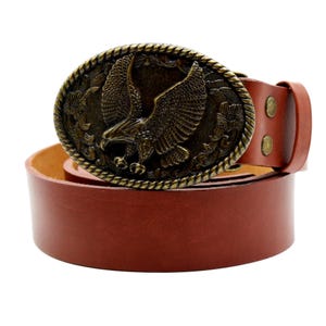 Puede incluir: Cinturón de cuero marrón con una hebilla ovalada de color bronce con un diseño de águila. La hebilla tiene un relieve detallado del águila con las alas extendidas, rodeada de motivos florales. El cinturón tiene un diseño clásico.