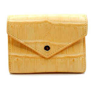 Puede incluir: Cartera de color amarillo claro con un patrón texturizado similar a la piel de cocodrilo. Tiene un cierre de solapa con un botón marrón oscuro. Los bordes están cosidos con hilo en contraste. La cartera es rectangular y parece ser de cuero.