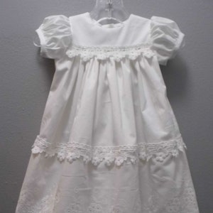 Puede incluir: Vestido de bautizo blanco con ribete de encaje y bordes festoneados. El vestido tiene una falda plisada y mangas abullonadas cortas.