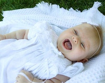 Puede incluir: Un vestido blanco de bebé con cuello de volantes y mangas cortas. El vestido está hecho de una tela suave y ligera y tiene un diseño simple y elegante. El bebé lleva una diadema blanca con un lazo.