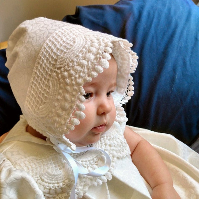 May include: A white baby bonnet with lace trim and a white satin ribbon. The bonnet is worn by a baby with a white dress.