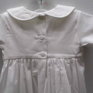 May include: White baby christening gown with a peter pan collar and short sleeves. The gown has a gathered skirt and is fastened with buttons down the back.