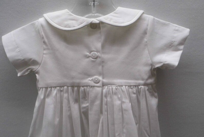 May include: White baby christening gown with a peter pan collar and short sleeves. The gown has a gathered skirt and is fastened with buttons down the back.