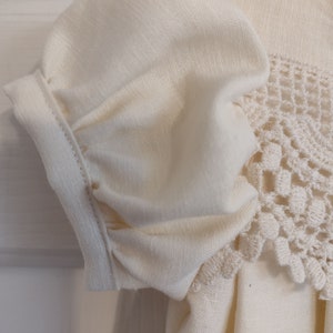 May include: A white cotton dress with a crocheted lace trim. The dress has a gathered puff sleeve and a simple, classic design.