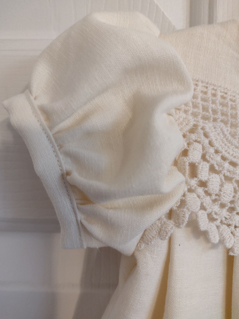 May include: A white cotton dress with a crocheted lace trim. The dress has a gathered puff sleeve and a simple, classic design.