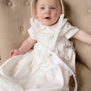 May include: A white baby bonnet with a white ribbon tied under the chin. The bonnet is worn by a baby sitting in a chair. The baby is wearing a white gown with a white ribbon tied around the waist.