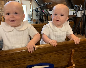 May include: Two babies in white outfits are sitting on a wooden pew in a church. They are both looking at the camera and smiling.
