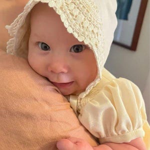 May include: A baby wearing a white crocheted bonnet with a lace trim and a white dress with puffed sleeves.