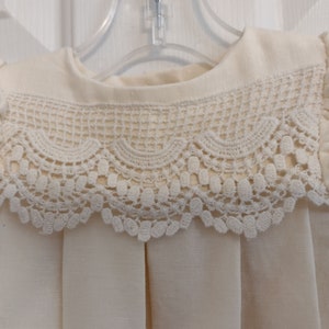 May include: A white baby dress with a lace collar. The dress has a round neckline and short puffed sleeves.
