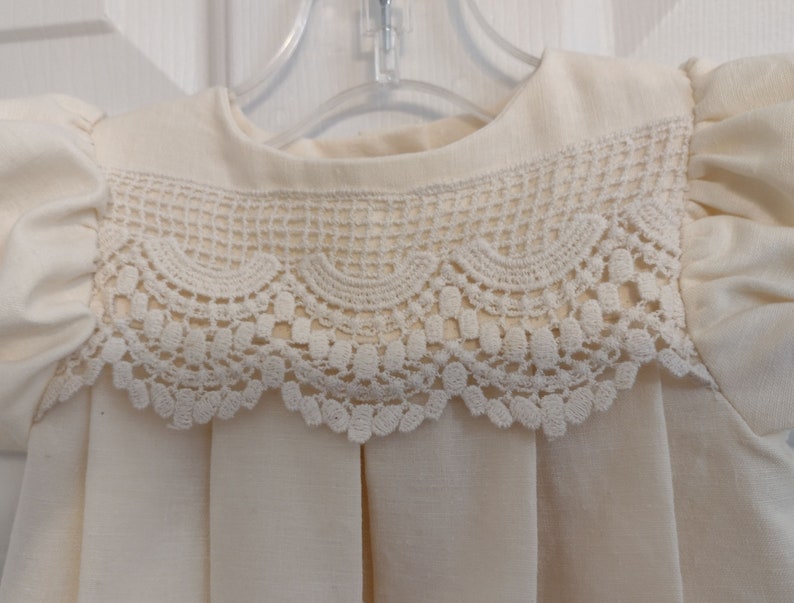 May include: A white baby dress with a lace collar. The dress has a round neckline and short puffed sleeves.