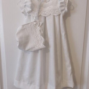 May include: White baby dress with a lace collar and matching bonnet. The dress has short sleeves and a gathered skirt.
