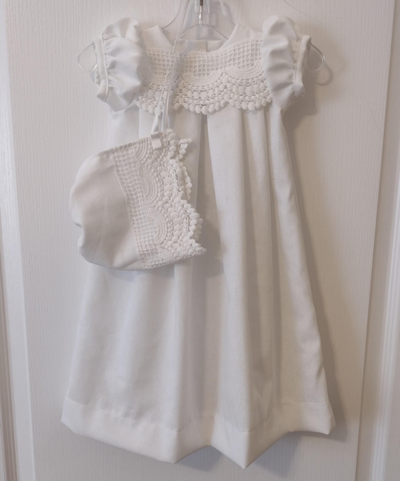 May include: White baby dress with a lace collar and matching bonnet. The dress has short sleeves and a gathered skirt.